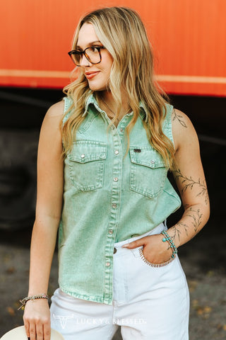 Woman wearing a green sleeveless denim shirt and white pants against an orange background