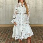 Woman in a white lace dress and brown boots standing against a metallic wall.