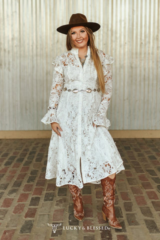 Woman in a white lace dress and brown boots standing against a metallic wall.