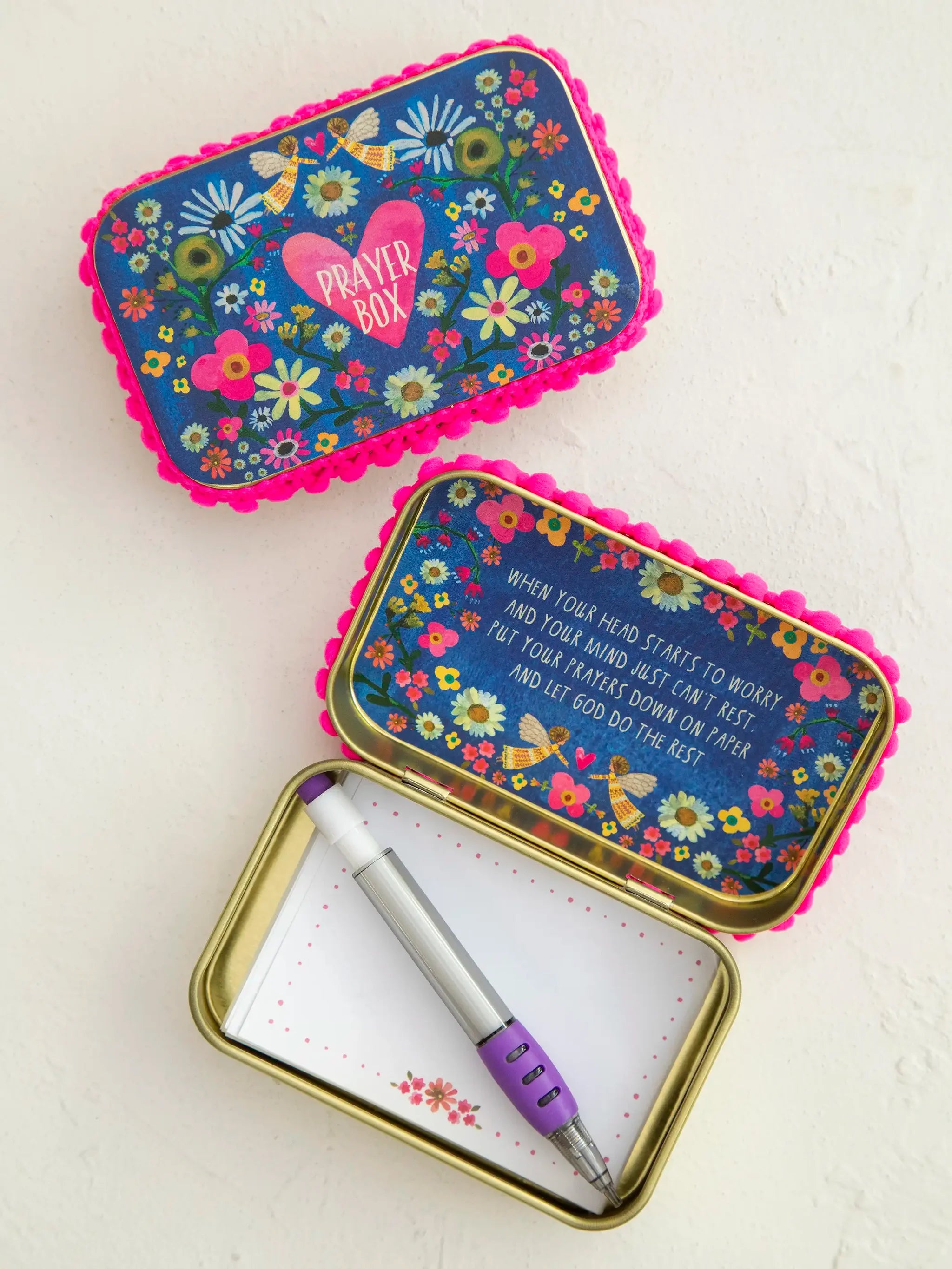 Two decorative metal prayer tin notebooks on a textured white background. One is closed, showing a blue floral lid with pink scalloped edges, a pink heart, and the words 'PRAYER BOX'.