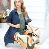A woman seated on a step wearing multicolored linen A-line pants and a navy blue shirt with a denim jacket draped over the shoulders.