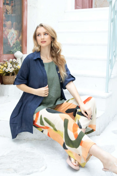 A woman seated on a step wearing multicolored linen A-line pants and a navy blue shirt with a denim jacket draped over the shoulders.