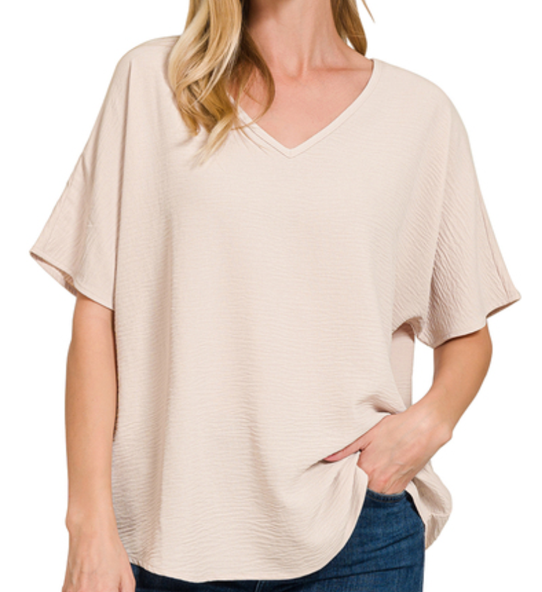 A woman wearing a light beige short-sleeve V-neck top with a textured crinkle fabric, paired with blue jeans, shown against a plain white background.