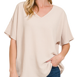 A woman wearing a light beige short-sleeve V-neck top with a textured crinkle fabric, paired with blue jeans, shown against a plain white background.