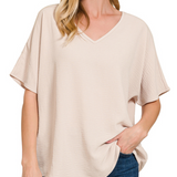 A woman wearing a light beige short-sleeve V-neck top with a textured crinkle fabric, paired with blue jeans, shown against a plain white background.