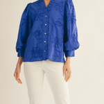 Woman wearing a blue embroidered blouse and white pants on a beige background