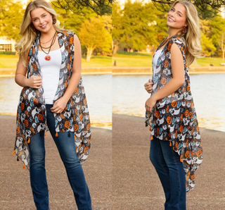 A woman modeling a sleeveless high-low open-front vest outdoors by a lake. The vest has a Halloween-themed print with orange pumpkins, white ghosts, and gray/black crows on a light gray background, and small orange tassels along the hem.