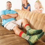 Man sitting on a couch wearing socks with designs and text, two children watching him.