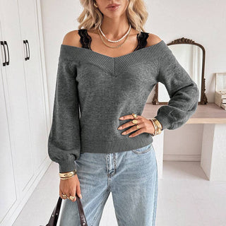 A woman wearing a dark gray off-the-shoulder knit sweater with a V-neckline, paired with light blue jeans and gold jewelry, standing in a bright, modern room.