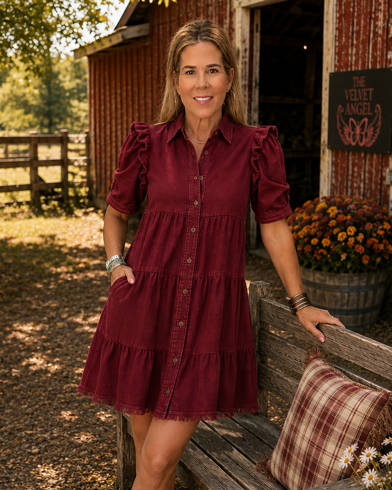 A woman wearing a tiered burgundy puff-sleeve button-up dress and tan cowboy boots stands next to a rustic wooden bench in front of a red barn, with flowers and a plaid pillow nearby.