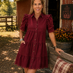 A woman wearing a tiered burgundy puff-sleeve button-up dress and tan cowboy boots stands next to a rustic wooden bench in front of a red barn, with flowers and a plaid pillow nearby.