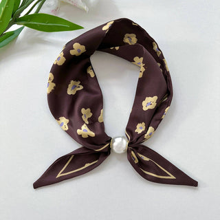 A dark brown silk scarf with a yellow floral pattern and a white faux-pearl knot, styled on a light gray surface with green leaves in the corner.