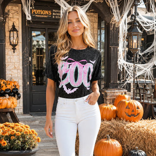 A woman stands outdoors in front of a Halloween-themed storefront, wearing a black graphic T-shirt with a large pink and white 'BOO' design and white pants.