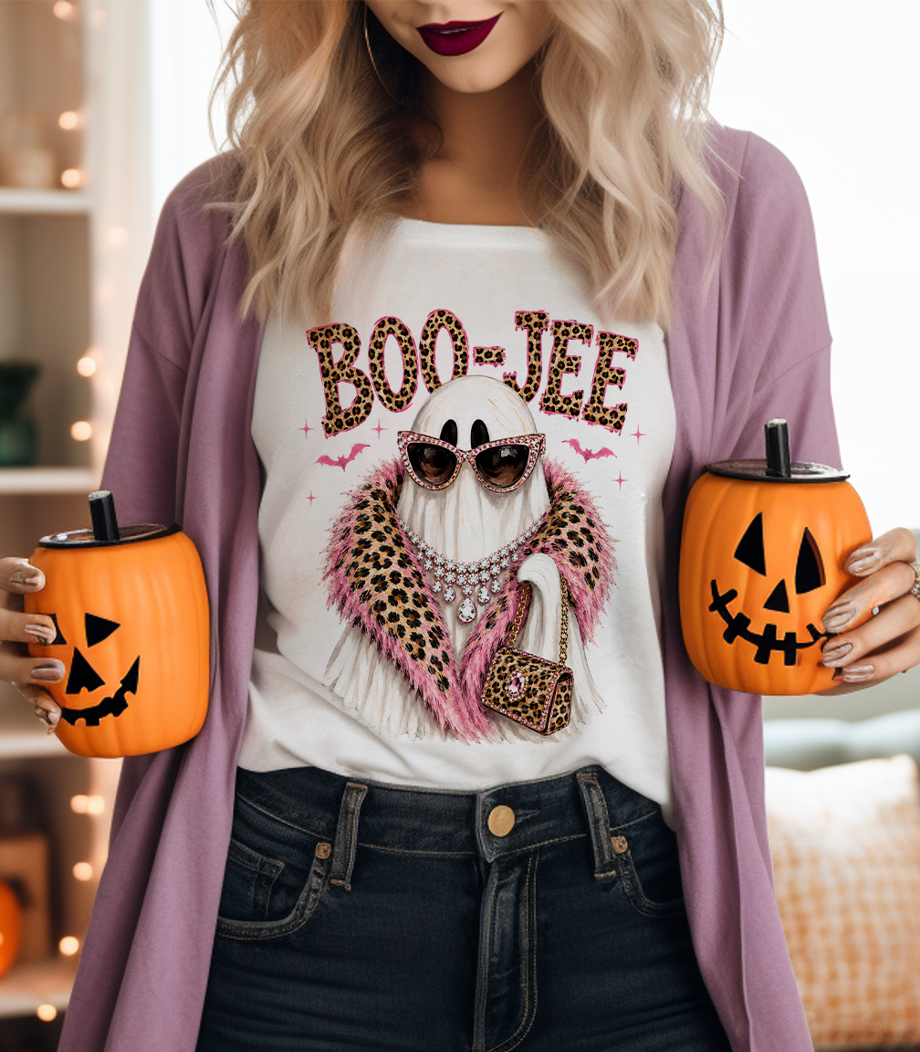 A woman wearing a white graphic T-shirt with a Halloween ghost design and a mauve cardigan, holding two orange jack-o'-lantern-shaped tumblers with black lids in a softly lit indoor setting.