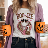 A woman wearing a white graphic T-shirt with a Halloween ghost design and a mauve cardigan, holding two orange jack-o'-lantern-shaped tumblers with black lids in a softly lit indoor setting.