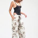 A woman modeling loose-fit jogger pants with an allover cowboy and cattle print in brown and beige on an off-white background, paired with a black camisole and white sneakers against a light gray studio backdrop.