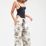 A woman modeling loose-fit jogger pants with an allover cowboy and cattle print in brown and beige on an off-white background, paired with a black camisole and white sneakers against a light gray studio backdrop.