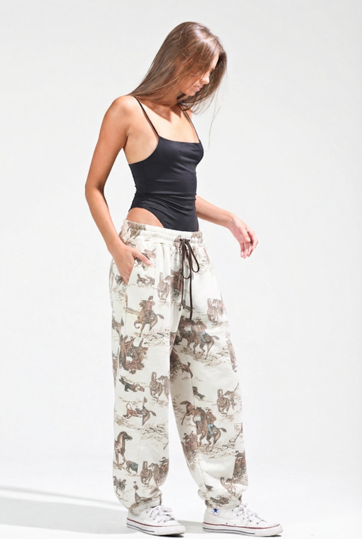 A woman modeling loose-fit jogger pants with an allover cowboy and cattle print in brown and beige on an off-white background, paired with a black camisole and white sneakers against a light gray studio backdrop.