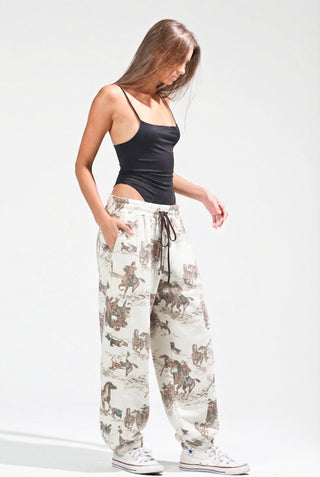 A woman modeling loose-fit jogger pants with an allover cowboy and cattle print in brown and beige on an off-white background, paired with a black camisole and white sneakers against a light gray studio backdrop.