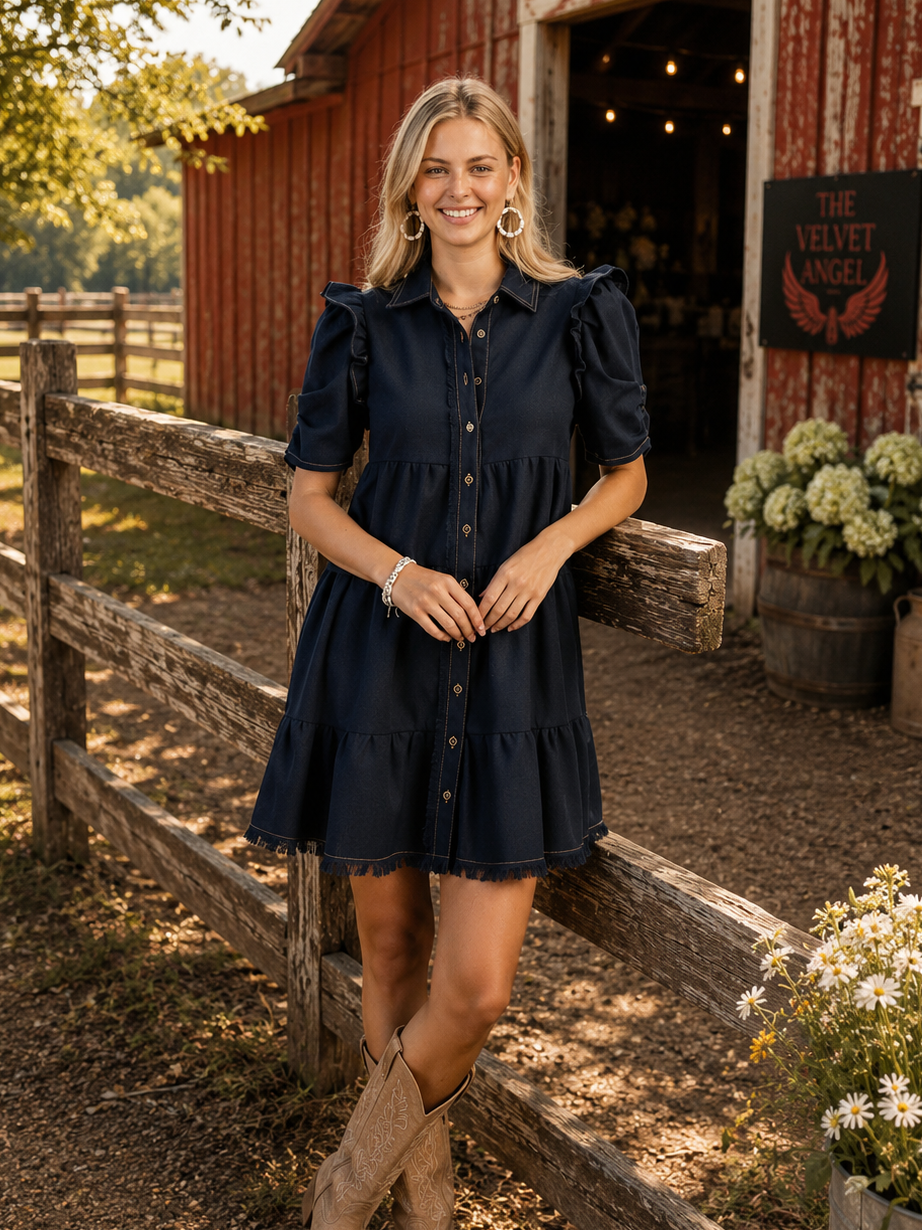 A woman stands outdoors in front of a red barn and wooden fence, wearing a short-sleeve navy blue button-front dress with ruffled shoulders and beige cowboy boots.