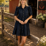 A woman stands outdoors in front of a red barn and wooden fence, wearing a short-sleeve navy blue button-front dress with ruffled shoulders and beige cowboy boots.