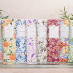 Set of colorful soap bars with decorative packaging on a wooden surface, surrounded by greenery.