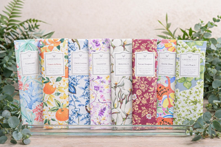 Set of colorful soap bars with decorative packaging on a wooden surface, surrounded by greenery.