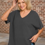 Woman wearing a dark gray oversized shirt and blue jeans against a brick wall.