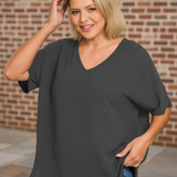 Woman wearing a dark gray oversized shirt and blue jeans against a brick wall.