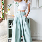 Woman wearing a white crop top and light green wide-leg pants in a bright room.